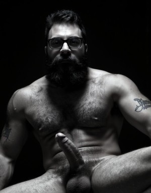 Fotografía hiperrealista en blanco y negro de un hombre adulto musculoso a la vista desde las rodillas para arriba hasta la cabeza, cuerpo muy delgado con musculación marcada y definida, rostro masculino, totalmente desnudo con polla erecta de treinta centímetros de longitud y ocho centímetros de diámetro, tiene rasgos marcados duros expresivos, barba densa poblada tupida oscura estilo hipster cubriendo completamente mandíbula mentón mejillas con grosor uniforme textura abundante muy definida, bigote denso tupido oscuro conectado perfectamente con la barba completa cubriendo completamente el labio superior, pelo oscuro medio peinado completamente recto hacia arriba con volumen considerable estilo hipster moderno quiff puntas verticales texturizadas hacia el cielo con altura prominente, cejas gruesas oscuras pobladas muy marcadas definidas, gafas de montura completa negra cuadradas grandes estilo hipster moderno sobre el puente de la nariz enmarcando unos ojos oscuros intensos que miran directamente a cámara con expresión seria penetrante confiada, expresión facial seria masculina sin sonrisa con mirada directa desafiante, pómulos marcados definidos angulares, nariz recta proporcionada, labios medios apenas visibles completamente cubiertos por bigote denso que conecta con la barba hipster completa, torso desnudo muy musculoso con pectorales enormes desarrollados y definidos, hombros anchos musculosos deltoide prominentes, brazos musculosos gruesos con bíceps y tríceps marcados visibles a ambos lados del cuerpo, abdomen tonificado marcado visible desde la cintura, piel con textura realista detallada poros imperfecciones sutiles, vello corporal denso oscuro abundante cubriendo pectorales abdomen brazos hombros con textura natural masculina muy visible, tatuaje en tinta negra en el bíceps del brazo derecho con la palabra exacta 