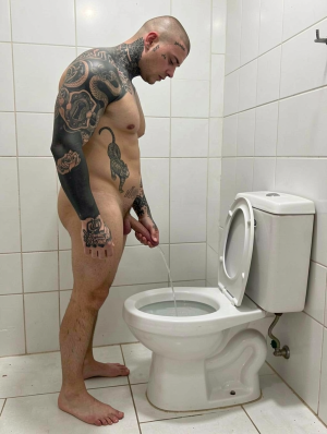 A naked, tattooed man is standing in a white-tiled bathroom, urinating directly into a toilet with the lid up. His body is shown in profile, with his shaved head tilted down as he watches the stream of urine falling into the bowl. He has numerous black and gray tattoos on his neck, chest, abdomen, arms, and one leg, including ornamental designs and large figures that cover much of his skin. The environment is simple and clinical, with a tiled floor, white walls, visible cistern, exposed pipes, and cold lighting that highlights the intimate, everyday scene.