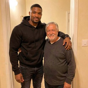 Two men pose together in a domestic interior, standing next to a door and beige-toned walls lit with warm light. On the left appears a tall, corpulent man with dark skin, short hair, and a trimmed beard, dressed in a black hooded sweatshirt and dark jeans; he smiles softly while putting his arm around the shoulders of the man beside him. On the right, an older, shorter man with gray hair combed back, a white beard, and a kind expression wears a dark gray sweater and dark pants. The scene conveys closeness, familiarity, and affection, like an informal keepsake photograph at home.