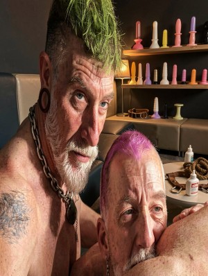Two older men with punk and fetish aesthetic pose very close in an intimate environment like a dungeon or sex room, with bare torsos, gauge piercings in their ears, thick chains around their necks and dyed mohawks, one green and the other pink. The man on the left stares fixedly at the camera with an intense expression, white beard and tattoo on his shoulder, while the other appears lower, with his face pressed against the bare skin of another person in the foreground, in an explicitly sensual pose. In the background there are shelves with dildos and sex toys of different sizes and colors, as well as lubricants, accessories and dark padded furniture, reinforcing a BDSM, queer and provocative atmosphere.