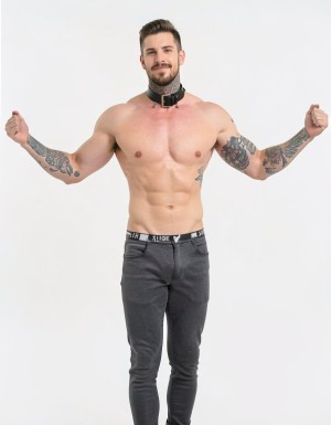 Hombre musculoso y sin camiseta posando de pie sobre un fondo blanco de estudio, con los brazos levantados y los puños cerrados en una actitud segura y desafiante. Luce un collar negro ancho de cuero con hebilla metálica alrededor del cuello, barba recortada, pelo corto peinado hacia arriba y una ligera sonrisa. Su torso definido deja visibles pectorales, abdominales y varios tatuajes en los brazos, hombros y costado. Viste pantalones ajustados de color gris oscuro, con la cinturilla de la ropa interior negra asomando por encima, creando una estética de moda masculina, fitness y estilo alternativo con aire dominante.