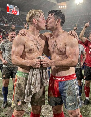 Dos futbolistas embarrados y sin camiseta se besan en mitad del campo, abrazados por los hombros tras un partido bajo la lluvia o sobre un césped muy deteriorado. El jugador de la izquierda, rubio y empapado, lleva pantalón corto manchado con el escudo del Sevilla y sostiene una camiseta o toalla sucia entre las manos; el de la derecha, moreno, también cubierto de barro y sudor, viste pantalón rojo y azul con la cintura visible. Alrededor, varios compañeros celebran eufóricos, algunos señalando hacia arriba, mientras el estadio lleno, los focos intensos y una pantalla gigante al fondo refuerzan el ambiente de victoria, celebración deportiva, fútbol, vestuario y emoción colectiva.