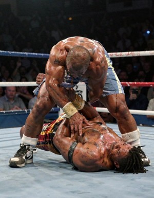 Dos boxeadores semi desnudos, muy musculosos, están luchando sobre el ring en el suelo, su piel  brilla por el sudor, destacan sus hombros, brazos y pectorales manchados de aceite, uno  de los rivales ha conseguido someter al otro, que yace KO, noqueado en el suelo 