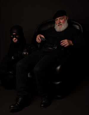 Un hombre mayor con barba blanca y gorra negra aparece sentado con actitud dominante en un sillón de cuero negro, vestido completamente de negro con jersey, pantalón, cinturón y botas, mientras sujeta una cadena o correa metálica con una mano. A su lado, en una posición baja y sumisa sobre el suelo, hay una persona enfundada en ropa negra brillante de cuero o látex, con pasamontañas ajustado que deja visibles los ojos y la boca. La escena tiene una iluminación tenue y dramática, fondo oscuro y estética fetichista, con un ambiente de poder, control, BDSM y retrato teatral de alto contraste.