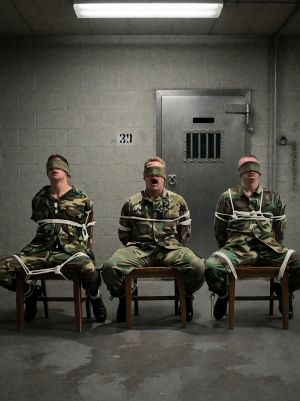 Tres hombres con uniforme militar de camuflaje permanecen sentados en sillas de madera dentro de una sala gris de aspecto carcelario, atados con cuerdas blancas alrededor del torso, los brazos y las piernas, e inmovilizados contra los respaldos. Los tres llevan vendas cubriéndoles los ojos; el hombre del centro tiene la boca abierta, como si estuviera gritando o respirando con dificultad, mientras los otros dos mantienen la cabeza ligeramente inclinada. El entorno es frío y opresivo, con paredes de bloques de hormigón, suelo de cemento manchado, una puerta metálica pesada con mirilla de barrotes y el número 30 pintado en la pared. Una luz fluorescente en el techo ilumina la escena con un tono duro e industrial, reforzando la sensación de interrogatorio, cautiverio y tensión militar.