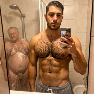 Un hombre joven, musculoso y con abundante vello en el pecho y el abdomen, se hace un selfi frente al espejo de un baño con azulejos beige, sujetando un móvil mientras lleva solo un pantalón corto gris de cintura elástica. Detrás de él, dentro de la ducha con mampara empañada y alcachofa visible, aparece un hombre mayor desnudo cubierto de espuma, con el vientre prominente, el pecho enjabonado y el pene parcialmente visible entre la espuma. La escena combina un retrato de gimnasio o físico masculino con un momento íntimo y cotidiano de ducha, iluminación cálida de baño, espejo, vapor y contraste generacional.