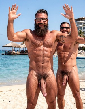 A highly detailed full-body nude photograph of a 40-year-old dominant muscle bear named Hipsterbarba, standing confidently in a well-lit studio with dramatic lighting that accentuates every muscle. He is 180cm tall, 80kg with 8% body fat, extremely muscular and symmetrical. Massive rounded hairy pecs with clear separation and dark pointed nipples, shredded six-pack abs with deep cuts and visible obliques forming a strong V-taper, wide shoulders, vascular arms, huge quads and defined legs. Very dense black masculine body hair covering his chest, running down in a thick treasure trail to a very bushy dark pubic area. He has a perfectly groomed long Bandholz beard that reaches mid-chest, thick mustache with curled upward spiral ends, modern high fade undercut hairstyle, black hipster glasses, and a small tattoo on his right bicep that reads 
