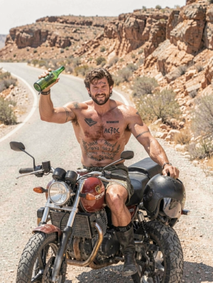 A shirtless man poses sitting on a classic Royal Enfield motorcycle on a desert road surrounded by rocky canyons, dry scrub, and an arid landscape in reddish tones. He is wearing green shorts, tall black boots, and multiple tattoos on his chest, arms, and torso, including the text AC/DC in the center; he has a beard, messy hair, and an expression somewhere between a smile and a grimace of effort under the intense sunlight. With one hand he raises a tilted green bottle, while the other rests near a black helmet placed on the motorcycle. The scene conveys a biker, adventurous, and wild style, with dust, a winding road, heat, and a desert-travel aesthetic.