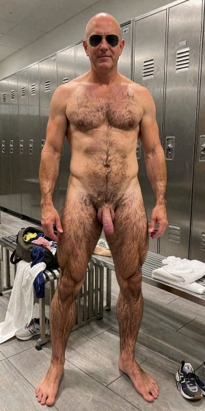 Bald and naked man standing in a locker room, facing forward with dark sunglasses and a straight, relaxed posture. He has a robust and hairy body, with abundant hair on his chest, abdomen, pubis, arms and legs; his penis and pubic hair are visible from the front. In the background there is a row of gray metal lockers with vents and locks, tiled floor and locker room benches. On the bench appear clothes, a white towel and personal items, while some sneakers are on the floor, reinforcing the context of a gym, shower or changing room.