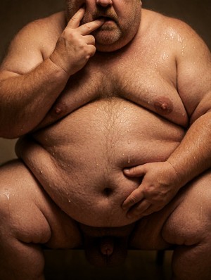 Un hombre desnudo de complexión muy obesa aparece sentado con las piernas abiertas, el torso y el abdomen cubiertos de gotas de agua que resaltan la piel húmeda, el vello corporal del pecho y la barriga prominente. La cabeza está parcialmente recortada, con barba corta y una mano apoyada en la cara, mientras la otra descansa sobre el vientre cerca de la ingle. Los pechos caen sobre el abdomen, los muslos ocupan gran parte de la escena y el pene queda visible entre las piernas. La iluminación cálida y el fondo oscuro crean un retrato corporal íntimo, crudo y frontal.