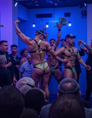 en un bar  con luz azul claro tres hombres musculosos vestidos con arneses, gorras y suspensorios bailan sobre el escenario con  erotismo desmedido, los clientes que son viejos en traje gritan e intentan tocarles,  les ofrecen billetes verdes que colocan  en sus tangas 