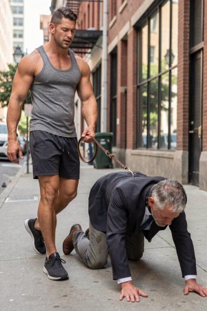 Un hombre musculoso con camiseta gris de tirantes, pantalón corto negro y zapatillas camina por una acera urbana sujetando una correa de cuero que va unida al cuello de otro hombre vestido con traje oscuro, camisa blanca y corbata, que avanza a cuatro patas sobre el pavimento. La escena transcurre entre edificios de ladrillo y grandes ventanales, con un ambiente de calle moderna y luz diurna. El contraste entre la postura dominante del hombre joven, erguido y atlético, y la posición sumisa del hombre canoso arrodillado crea una imagen de humillación pública, control, fetichismo y dinámica de poder tipo BDSM en un entorno cotidiano.