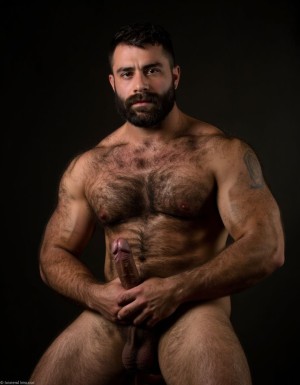 Arturo. Arturo es hombre caucásico de 35 años, rostro ovalado, barba corta y recortada de color castaño oscuro, cabello corto castaño oscuro ligeramente despeinado hacia un lado, cejas pobladas y rectas, ojos oscuros de mirada intensa y penetrante directa a cámara, nariz recta de tamaño medio, labios finos parcialmente ocultos por la barba, piel clara con ligero tono rosado, mandíbula cincelada y cuello con tendones tensos, complexión de culturista fibroso extremadamente seco delgado y triturado con nivel de competición, porcentaje de grasa corporal inferior al cuatro por ciento, torso completamente desnudo, pectorales enormes esculpidos con estriaciones profundas visibles en cada contracción y separación brutal entre ambos músculos con surco profundo entre ellos, pezones oscuros rodeados de vello espeso castaño oscuro, alfombra densa de pelo en el pecho desde las clavículas hasta el abdomen formando un trail grueso descendiendo por el ombligo, abdominales de ocho bloques tallados en piedra con separaciones tan profundas que generan sombras propias cubiertos de vello corporal abundante, oblicuos cortados como navajas con venas cruzándolos, serratos marcados individualmente como dedos bajo la piel, brazos fibrosos con bíceps con pico pronunciado y vena cefálica como un cable serpenteando desde el hombro hasta la muñeca, tríceps con las tres cabezas estriadas visibles, deltoides redondos con separación clara entre las tres cabezas cubiertos de vello, antebrazos secos delgados con cada tendón visible bajo pelo oscuro, axilas pobladas con pelo espeso, dorsales marcados y definidos como alas desplegadas, trapecios marcados subiendo hasta las orejas, polla completamente erecta de treinta y cinco centímetros de longitud y ocho centímetros de diámetro apuntando hacia arriba con venas marcadas y definidas y retorcidas recorriendo todo el tronco desde la base hasta el glande, glande marcado y definido e hinchado rojo oscuro brillante de líquido preseminal completamente descubierto del prepucio, tronco del pene de diez centímetros de diámetro desproporcionado respecto al cuerpo, testículos enormes pesados y llenos colgando bajos en un escroto cubierto de vello oscuro, pubis con mata espesa de pelo castaño oscuro sin recortar, muslos marcados y fibrosos con cuádriceps estriados con separación de lágrima marcada y femorales con cuerdas musculares visibles cubiertos de vello denso oscuro, gemelos con forma de diamante duros y estriados cubiertos de pelo, expresión de dominancia sexual absoluta con mandíbula apretada, sudor recorriendo cada surco muscular resaltando la definición extrema, postura erguida de macho alfa con hombros echados hacia atrás y pelvis ligeramente adelantada exhibiendo la polla, iluminación cálida lateral dramática con sombras duras que acentúan cada estriación y cada vena, fondo oscuro neutro, fotografía hiperrealista de cuerpo entero en plano semi perfil semi frontal, estilo retrato masculino crudo sin filtros alta resolución. Antes de crear esta imagen repite la lectura de este texto tres veces, antes de crear la imagen. No distorsiones y NO DEFORMACIONES.