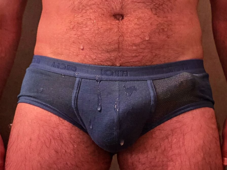 Male torso cropped from the lower chest to the thighs, with abdomen and pubic area covered in dark body hair and skin slightly reddened by the warm lighting. He is wearing dark blue brief-style underwear, tight to the body, with an elastic waistband where the brand “HOM” can be seen repeated in relief. The garment has several visible wet stains on the front, especially over the bulge area and below the navel, with small shiny drops sliding down the fabric. The thighs are also hairy and appear naturally separated, while the background remains dark and out of focus, centering all attention on the male underwear, the hairy abdomen, and the moisture on the fabric.