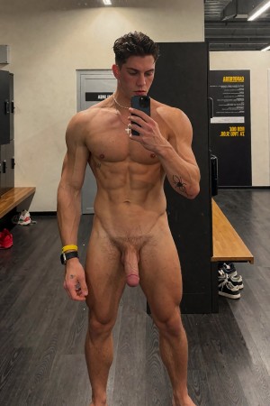 Un hombre joven y musculoso posa desnudo frente a un espejo de vestuario de gimnasio mientras se hace un selfi con el móvil, mirando la pantalla con expresión seria. Su torso definido, abdominales marcados, brazos fuertes y piernas atléticas destacan bajo la iluminación interior; lleva una cadena con cruz, una pulsera amarilla y un reloj o pulsera negra. El pene y el vello púbico son claramente visibles. Al fondo aparecen taquillas negras, bancos de madera, suelo oscuro, zapatillas en el suelo y carteles de normas del gimnasio, creando un ambiente de vestuario moderno y deportivo.