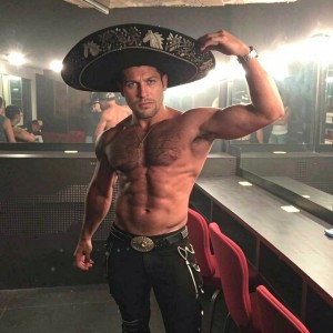 Hombre musculoso sin camiseta posa en un camerino iluminado, levantando un brazo para marcar el bíceps mientras sostiene un gran sombrero de mariachi negro con bordados plateados y motivos decorativos. Su torso definido, abdominales marcados y pecho velludo destacan bajo la luz directa de los espejos, creando reflejos alrededor de la estancia. Lleva pantalón negro ajustado con cinturón de hebilla grande y detalles metálicos, además de un colgante y reloj. El ambiente parece un backstage o vestuario, con tocadores, espejos, sillas rojas y encimeras largas, transmitiendo una mezcla de estética mexicana, pose de fisicoculturismo y preparación escénica.