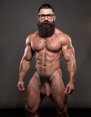 A highly detailed full-body nude photograph of a 40-year-old dominant muscle bear named Hipsterbarba, standing confidently in a well-lit studio with dramatic lighting that accentuates every muscle. He is 180cm tall, 80kg with 8% body fat, extremely muscular and symmetrical. Massive rounded hairy pecs with clear separation and dark pointed nipples, shredded six-pack abs with deep cuts and visible obliques forming a strong V-taper, wide shoulders, vascular arms, huge quads and defined legs. Very dense black masculine body hair covering his chest, running down in a thick treasure trail to a very bushy dark pubic area. He has a perfectly groomed long Bandholz beard that reaches mid-chest, thick mustache with curled upward spiral ends, modern high fade undercut hairstyle, black hipster glasses, and a small tattoo on his right bicep that reads 