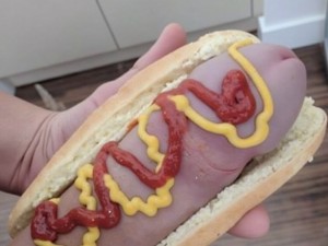 En una cocina vieja se ve un primer plano de una mano que sostiene un pan de perrito (hot dog) que tiene dentro una salchicha de color carne y con forma de pene, le han puesto mostaza, tomate ketchup, se muestran  los envases de tomate, mayonesa, mostaza y artículos de cocina en la encimera.