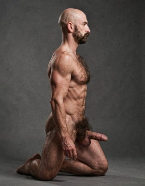 Naked man in profile kneeling on a gray studio floor, with his torso upright, shaved head and trimmed dark beard, looking to the right with a serious, neutral expression. His muscular body is lit from the side, highlighting shoulders, defined arms, hairy chest, sculpted abdomen, and veins in the forearm. The pose leaves the bent legs visible, the feet resting backward, abundant pubic hair, and the flaccid penis oriented forward. The plain, dark background, with a slightly rough texture, creates a sober and artistic composition centered on male anatomy, partial frontal nudity, and body study.