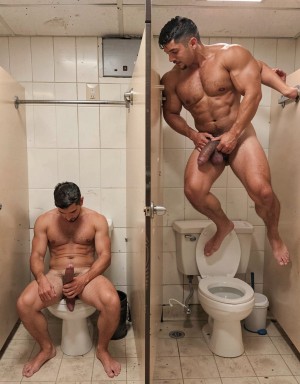 Dos hombres desnudos aparecen en un baño público de azulejos beige con cabinas separadas por paneles, inodoros, barras metálicas y suelo desgastado; a la izquierda, un hombre sentado en el váter mira hacia abajo mientras sostiene su pene erecto con una mano, con postura relajada y las piernas abiertas. A la derecha, un hombre muy musculado y depilado está elevado junto al inodoro, apoyado en la estructura de la cabina, con una pierna flexionada y el cuerpo inclinado hacia el centro, sujetándose el pene con la mano mientras mira hacia abajo. La escena tiene iluminación fría de baño, ambiente íntimo y explícito, con énfasis en desnudez masculina, musculatura marcada, urinarios privados e interacción sexual en un aseo público.