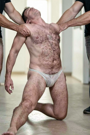 Un hombre con el torso desnudo y abundante vello corporal aparece arrodillado en un pasillo luminoso, con la cabeza echada hacia atrás y una expresión de tensión o abandono mientras dos personas vestidas de negro le sujetan con fuerza por los brazos y las axilas. Lleva únicamente un slip blanco translúcido de encaje o malla fina, muy ajustado y semitransparente, que marca claramente el pene bajo la tela. La postura forzada, una pierna extendida hacia delante y la otra doblada sobre el suelo, transmite una escena física intensa, casi performativa, con sensación de control, vulnerabilidad y sujeción corporal.