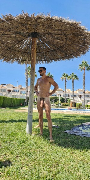 Un hombre desnudo posa de pie sobre el césped, bajo una sombrilla de paja tipo brezo que proyecta sombra en una zona ajardinada junto a una piscina. Lleva gafas de sol oscuras, barba corta y el pelo oscuro, con las manos apoyadas en la cintura y una postura relajada; su cuerpo está completamente al descubierto, incluido el pene y el vello púbico. Alrededor aparecen palmeras, setos cuidados, tumbonas y edificios blancos de estilo vacacional, creando un ambiente de resort o urbanización turística en un día soleado con cielo azul despejado.