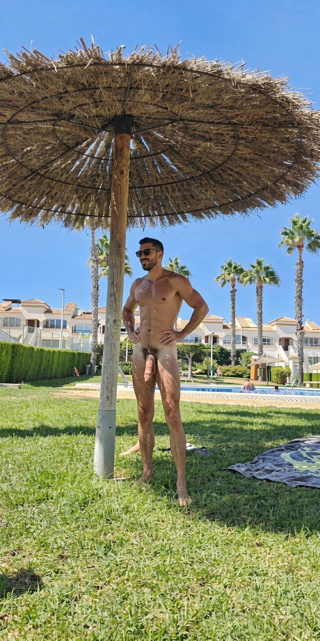 Un hombre desnudo posa de pie sobre el césped, bajo una sombrilla de paja tipo brezo que proyecta sombra en una zona ajardinada junto a una piscina. Lleva gafas de sol oscuras, barba corta y el pelo oscuro, con las manos apoyadas en la cintura y una postura relajada; su cuerpo está completamente al descubierto, incluido el pene y el vello púbico. Alrededor aparecen palmeras, setos cuidados, tumbonas y edificios blancos de estilo vacacional, creando un ambiente de resort o urbanización turística en un día soleado con cielo azul despejado.
