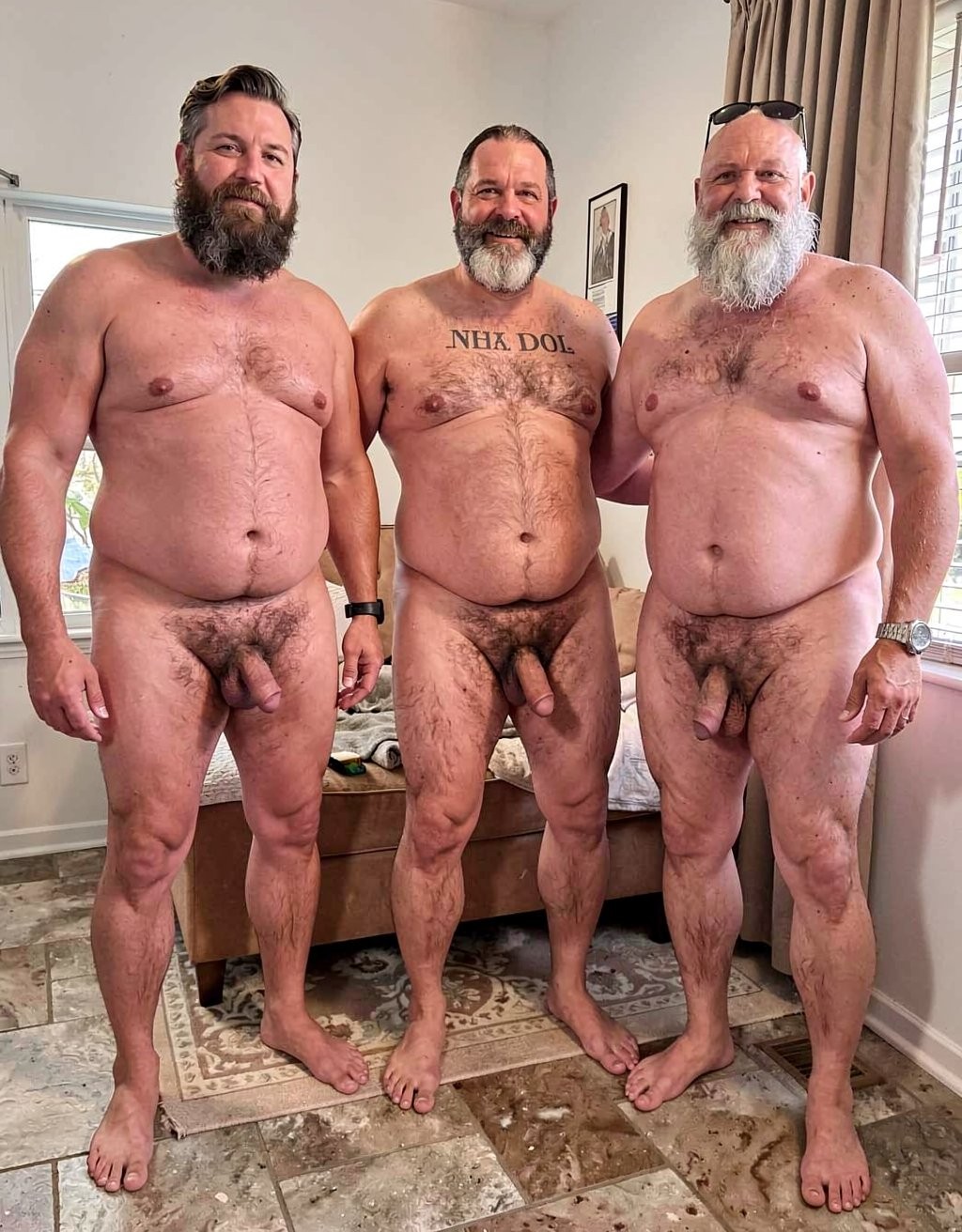 Three naked men pose standing in a bright room, looking at the camera with a relaxed and smiling attitude; all three have abundant beards, robust bodies, and visible body hair on their chest, abdomen, legs, and pubic area. Their penises and testicles are completely exposed in a frontal pose, with no sexual interaction, their arms close to or resting on each other like a group portrait. The man in the center has a text tattoo on his chest, while the one on the right has sunglasses on his head and a watch on his wrist. The setting appears domestic, with tiled flooring, a rug, a low bed or sofa, beige curtains, and a window with blinds that provides natural light.