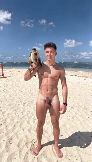 Hombre adulto desnudo de pie sobre una playa de arena blanca, sosteniendo un pequeño mono sobre el hombro izquierdo. Tiene el pelo oscuro peinado hacia arriba, complexión atlética y musculosa, y mira a cámara con una leve sonrisa; su pene y testículos quedan visibles. Lleva una pulsera negra en la muñeca derecha. Detrás se extiende el mar turquesa con pequeñas olas, varias embarcaciones lejanas y un cielo azul luminoso salpicado de nubes blancas, mientras su sombra se proyecta sobre la arena.