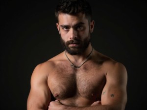 Hombre español dominante de 35 años, pelo castaño oscuro con barba poblada, mirada intensa y penetrante, torso musculoso desnudo, brazos cruzados, tatuajes en el pecho, expresión seria y autoritaria, iluminación dramática, fondo oscuro minimalista, cuerpo atlético definido, postura dominante y segura, piel mediterránea bronceada, mandíbula marcada.