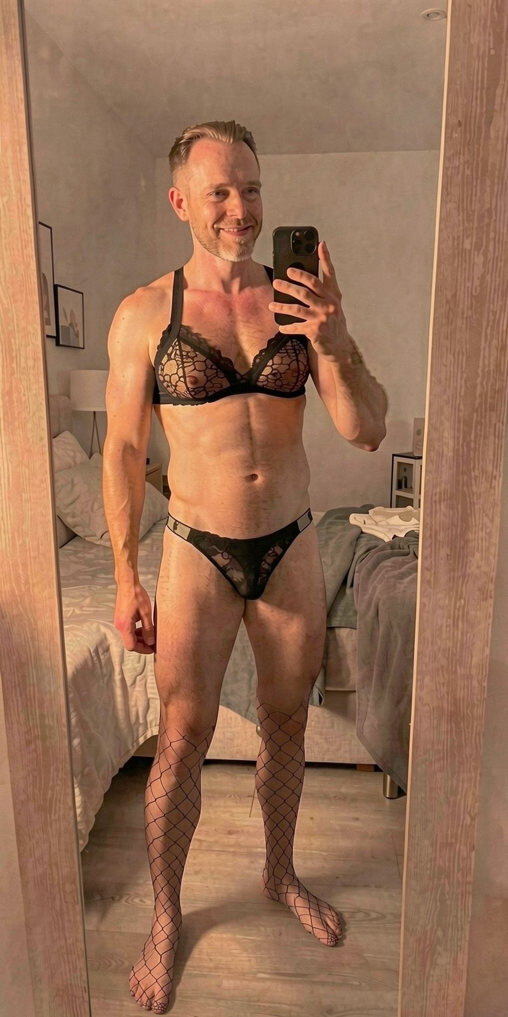 Hombre sonriente haciéndose un selfie frente a un espejo de cuerpo entero en un dormitorio, sujetando el móvil con una mano y posando con lencería negra de encaje. Lleva un sujetador triangular transparente con detalles florales, un tanga negro estrecho que cubre el pene y medias de rejilla que suben por las piernas, creando un estilo sensual y provocador. Su cuerpo atlético y depilado queda casi completamente expuesto, con brazos definidos y postura relajada. Al fondo aparece una cama deshecha con sábanas claras, una mesilla con lámpara y ropa sobre el borde, todo iluminado por una luz cálida e íntima.