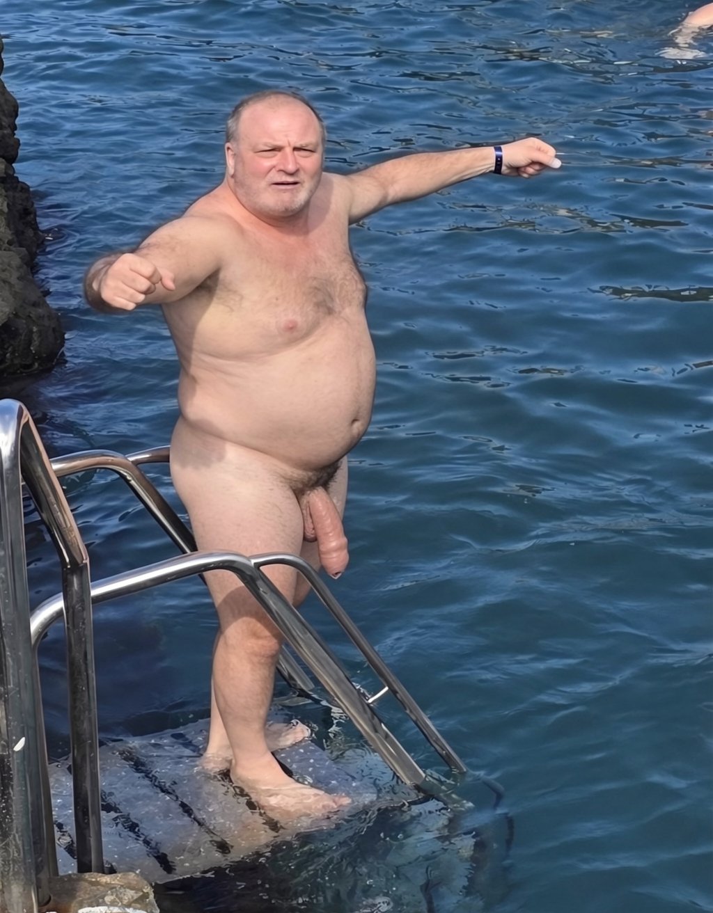 A naked man is standing on a small metal platform by the sea, holding onto the stainless steel bathing ladder while pointing both arms forward, with a serious or focused expression. The blue water surrounds the scene, and a dark rocky wall can be seen to one side, suggesting a coastal area or access to the sea from rocks. His body is completely exposed, with his torso, belly, legs, and penis visible, and he is wearing a dark bracelet or watch on one wrist. The sunlight illuminates his skin and creates reflections on the waves, giving the scene a spontaneous open-air bathing appearance in the sea.