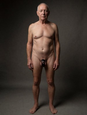 Elderly naked man standing, photographed full body in a studio with a dark gray background and soft side lighting that highlights the texture of the skin, torso, arms, and legs. He is in a frontal posture, upright, with arms relaxed at both sides and bare feet slightly apart, looking directly at the camera with a serious and neutral expression. He has very short gray hair, a slim build, and a bare chest; the genital area appears partially covered by a tight black device, similar to a protector or harness, placed over the penis. The composition is sober, minimalist, and an artistic portrait, centered on the human body, old age, male nudity, and physical presence without adornments.