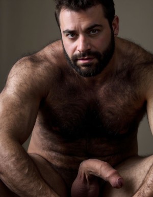 Hombre con mucho vello corporal musculoso totalmente desnudo agachado con pene erecto.   Cara con hoyuelo en barbilla, mentón hendido, el mentón está limpio y bien afeitado, NO BARBA y NO BIGOTE, cara masculina de un hombre totalmente desnudo de cuerpo entero y muy viril y musculoso y guapo y sexi de 40 años que tiene mandíbula fuerte y cuadrada, destacando una fisura vertical marcada en el centro del mentón que es el hoyuelo. La cara es de un hombre español musculoso culturista, torso desnudo, constitución corporal extremadamente desarrollada escultural, boca cerrada en sonrisa torcida arrogante burlona, labios carnosos, ojos verdes claro profundos con mirada penetrante directa a cámara cargada de cinismo y dominancia, cejas pobladas densas, cabello denso despeinado hacia atrás en copete desordenado con mechones rebeldes cayendo sobre frente y sienes mostrando textura natural sin producto, cuello musculoso con nuez prominente visible, trapecios enormes elevados como montañas a ambos lados del cuello, pecho descomunal con pectorales gigantescos separados con hendidura profunda central cada fibra muscular visible bajo piel bronceada cubiertos completamente por vello denso o negro y tupido salvaje sobre toda la superficie con pezones oscuros erectos asomando entre pelaje espeso, abdomen perfectamente esculpido en tableta de ocho secciones profundas con sombras dramáticas entre cada músculo, oblicuos cortados laterales, línea de vello negro denso descendiendo desde centro del pecho entre pectorales por esternón sobre cada abdominal hasta ombligo donde se ensancha formando trail oscuro salvaje continuando descendiendo, brazos con bíceps de gran tamaño con cabezas musculares divididas y venas gruesas azules serpenteando por toda superficie, tríceps marcados en herradura, deltoides esféricos estriados, antebrazos masivos cubiertos de vello negro denso con venas prominentes bajando hasta muñecas, dorsales anchos como alas creando V invertida brutal, piel clara ligeramente bronceada con poros visibles micro imperfecciones naturales lunares y marcas sutiles, vello corporal negro abundante cubriendo completamente pecho abdomen brazos antebrazos cuello en densidad natural masculina sin recortar, iluminación natural lateral dramática creando sombras duras acentuando cada músculo, fondo neutro desenfocado, estilo fotografía editorial cruda realista, grano fotográfico sutil, colores naturales intensos saturados. Está totalmente desnudo