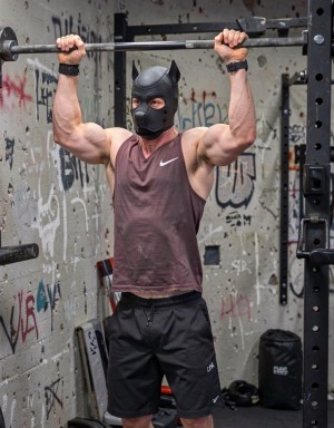 Shirtless man wearing a black puppy hood. He is in a gritty gym, doing pullups, standing and his hands gripping a steel bar above him.
