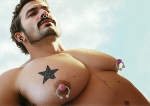 hombre de mediana edad que muestra grandes pectorales sin vello, tiene dos piercings de herradura en sus grandes pezones, así como en la nariz y oreja. Lleva cabelllo un poco largo, bigote prominente y barba de días. 