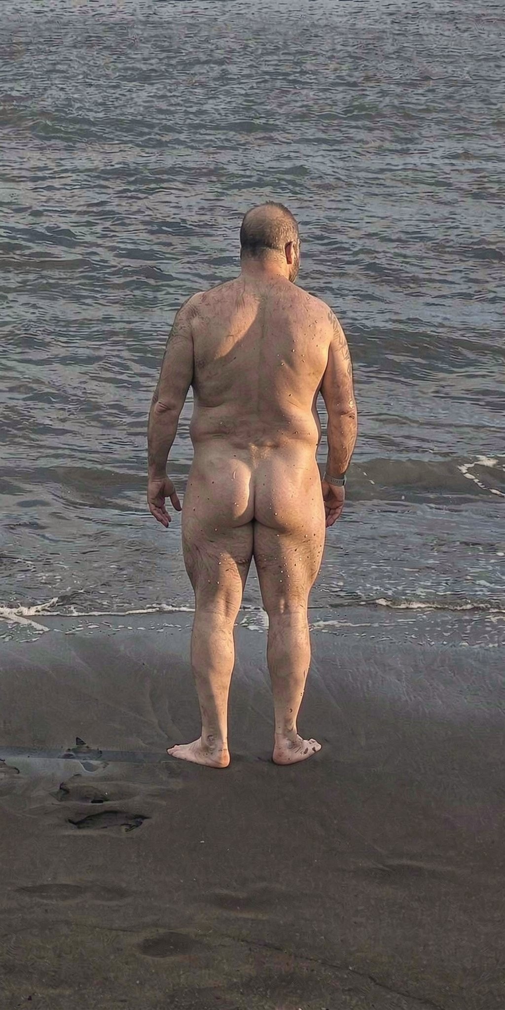 A naked man, seen from behind, stands on the shore of a dark sandy beach while looking out to sea, with his feet near the foam of the waves. His body is completely exposed from behind, with his back, buttocks, and legs visible, and the light highlights the texture of his skin and natural musculature. The water fills the background with small gray and green ripples, creating a calm coastal atmosphere. Footprints and remnants of foam appear in the wet sand, reinforcing the feeling of a spontaneous scene by the sea.