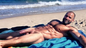 Fotografía panorámica 16:9 en ultra alta definición para imágenes 4K: hombre de 35 años de edad está acostado sobre una toalla en la playa, es delgado, músculos muy definidos y fibrosos delgado, totalmente desnudo con mucho vello por todo el cuerpo, se ven sus testículos hermosos y con mucho vello, cuerpo sudoroso y bronceado. Tiene un hermoso pene que está erecto y es acariciado vigorosamente con la mano derecha.