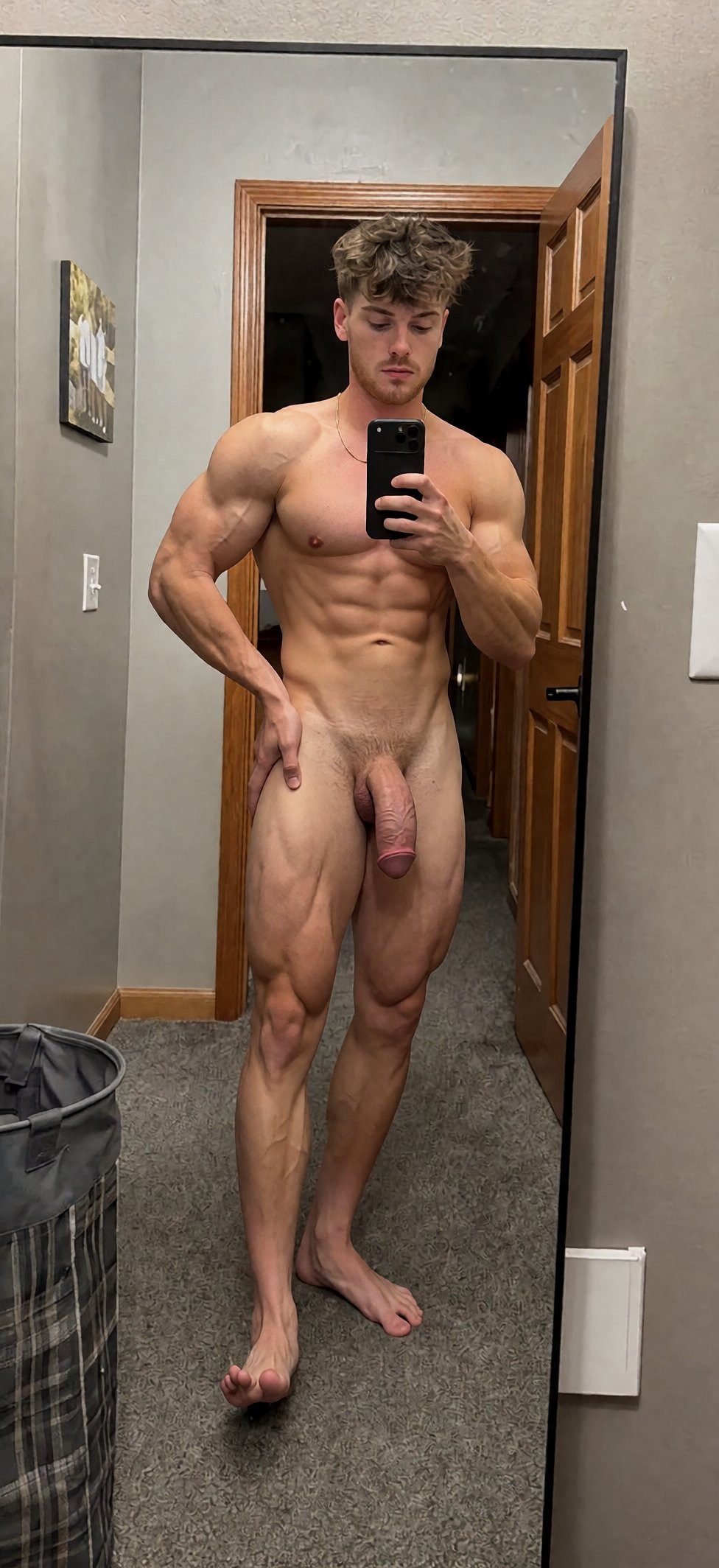 Hombre desnudo y muy musculado posando frente a un espejo de cuerpo entero mientras se hace un selfie con el móvil, en una habitación doméstica con paredes grises, moqueta y una puerta de madera abierta al fondo. Su físico destaca por los brazos voluminosos, pectorales marcados, abdominales definidos y piernas muy desarrolladas, con venas visibles y postura de culturismo. Lleva el pelo castaño despeinado y mira la pantalla del teléfono con expresión neutra; su pene cuelga visible entre los muslos. A un lado aparece un cesto de ropa y en la pared se aprecia un pequeño cuadro, reforzando el ambiente íntimo de dormitorio o vestidor.