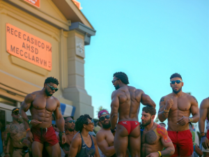 Orgullo madrileño a su paso por la puerta de Alcalá, se muestra la parte alta del monumento. Varios negros y mulatos musculosos subidos en la carroza, muestran sus cuerpos y uno de ellos está en tanga con un culazo, el que está a su lado le toca el paquete. Hay gente por todas partes y subida en la carroza. Luz del atardecer reflejada sobre el monumento.
