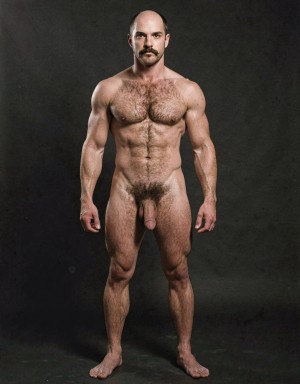 Mike Morris, a 32-year-old Caucasian male with an extremely lean and athletic physique at only 8% body fat, stands completely naked in a strict military attention pose. Full body frontal shot from the tips of his toes to the top of his head. His body is young, dry, and highly defined with sharp muscle separation, visible vascularity, and a very narrow waist. He has advanced Norwood III-IV male pattern baldness with a shiny tanned scalp on top and only short dark brown hair on the sides and back. His face is serious and commanding: square angular jaw, prominent cheekbones, thick dark handlebar mustache, heavy brows, and intense deep-set brown eyes. His massive square pectorals are pushed forward, covered in dense dark brown chest hair that forms a classic thick T-pattern running down the sternum and spreading over his sharply carved six-pack abs. Small dark nipples sit low on the pecs. A prominent trail of dark hair leads to an extremely dense, curly, dark brown pubic bush that climbs almost to his navel. His thick 14 cm flaccid cock hangs heavy over large, low-hanging, hairy testicles, the shaft skin noticeably lighter than his bronzed body. Powerful veined forearms, developed traps, defined quadriceps with clear separation, diamond-shaped calves, and slightly hairy feet complete his figure. He stands perfectly straight, chest out, shoulders back, arms rigid at his sides with fists clenched, head high and gaze fixed forward. Dramatic directional studio lighting from above and slightly to the front creates strong highlights on his dry, vascular skin and deep shadows that accentuate every muscle fiber, the texture of his dense body hair, and the extreme definition of his 8% body fat physique. Photorealistic, high-contrast, 1970s Colt Studio masculine aesthetic, grainy film texture. Tiene un bañador verde oscuro de piscina.