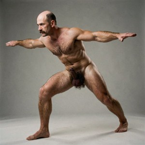 Mike Morris, a 32-year-old extremely lean and shredded Caucasian male with only 8% body fat, stands completely naked in a perfect Warrior III yoga pose, captured in a full uninterrupted body shot from the tips of his grounded foot all the way to the top of his bald head and the tips of his extended arms and lifted foot. The composition is taken from a three-quarter angle with a slight 45-degree foreshortening that adds depth and power to the entire figure. He balances powerfully on one straight leg, the other leg extended perfectly straight back parallel to the floor, torso hinged forward at the hips forming a clean horizontal line from head to lifted foot, arms stretched forward aligned with the body. The 45-degree three-quarter view reveals the dramatic depth of his V-tapered back, rounded and very hairy glutes, the full side profile of his deeply etched six-pack abdominals and obliques, massive squared pectorals, thick vascular thighs and diamond-shaped calves. His entire physique is covered in dense uniform dark brown body hair that forms a thick pattern across his chest, shoulders, arms, abdomen and lower back, exploding into an extremely dense, curly pubic bush that climbs almost to his navel. Dramatic studio lighting falls from high and slightly to the side at 45 degrees, creating sharp rim lighting that traces every muscular contour, every vein and every striation while casting deep, rich shadows that emphasize the extreme dryness, vascularity and three-dimensional form of his body. His head is bald on top with very short dark brown hair on the sides and nape, hard angular face visible in three-quarter view with square jaw, thick well-groomed handlebar mustache and intense dark brown eyes looking forward with focused expression. His genitals hang naturally and completely flaccid between his legs in this angled view: a thick soft 14 cm cock with long foreskin fully covering the glans, visible veins and noticeably lighter skin tone that contrasts sharply with his bronzed hairy torso, paired with large, heavy, low-hanging testicles with wrinkled darker skin, all framed by the thick dark brown curly pubic hair. Every anatomical detail from the hairy toes on his size 44 feet, powerful muscular legs, contracted abs, veined forearms, rounded hairy glutes, soft hanging cock and balls to the perfect straight-line tension of the Warrior III pose is rendered with absolute precision. Photorealistic, high-contrast, masculine 1970s Colt Studio aesthetic with subtle grainy film texture and masterful dramatic studio lighting.
