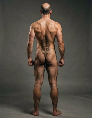 Mike Morris, a 32-year-old extremely lean Caucasian male at only 8% body fat, stands completely naked in a strict military attention pose seen from directly behind in a full posterior view. His back is perfectly straight, shoulders pulled back, arms rigid at his sides with fists clenched, legs straight and together, head held high. The shot captures him from the heels of his feet all the way up to the top of his bald head. His advanced Norwood III-IV baldness is clearly visible from behind, with a smooth tanned scalp on the crown and short dense dark brown hair only on the sides and lower back of the head. His back is highly defined and vascular: wide lats tapering dramatically to a very narrow, tight waist, thick trapezius muscles, and dense dark brown body hair lightly covering the shoulders and becoming much thicker and denser across the lower back and lumbar area, especially between the lats. His ass is tight, muscular and round with almost no fat, the glutes clearly separated. Thick dark hair continues down onto his powerful diamond-shaped calves and the backs of his slightly hairy feet. Dramatic directional studio lighting from above and slightly to the side creates strong highlights on his dry, shredded skin and deep shadows that accentuate every muscle fiber, the texture of his dense back hair, and the extreme vascularity and definition of his 8% body fat physique. Photorealistic, high-contrast, masculine 1970s Colt Studio aesthetic, grainy film texture, commanding and powerful presence.