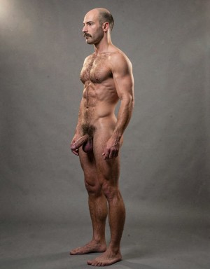 Mike Morris, a 32-year-old extremely lean Caucasian male with only 8% body fat, stands completely naked in a strict military attention pose captured in a full-body exact side profile at 90 degrees from his left side. The shot runs from the tips of his size 44 slightly hairy feet all the way up to the top of his bald head, showing his entire body without any cropping. His physique is dry, highly vascular and powerfully defined: massive square pectorals seen in perfect side silhouette, dense dark brown chest hair forming a thick T-pattern, sharply carved six-pack abs in clear profile, narrow waist, wide lats, powerful thighs and diamond-shaped calves. Dramatic directional studio lighting comes from high and slightly in front of him, creating strong rim lighting that traces the sharp outline of his entire body, the texture of his dense dark body hair on chest, lower back, ass and legs, and deep shadows that accentuate every muscle separation, vein and the extreme dryness of his 8% body fat physique. His advanced Norwood III-IV bald scalp shines under the light, with short dense dark brown hair on the sides and back of his head. His square angular face is shown in pure profile: strong jawline, prominent chin with slight central dimple, thick dark handlebar mustache, heavy brow and intense deep-set brown eye looking straight forward with a serious, commanding expression. His penis is fully visible in perfect side view, completely natural and realistic: thick, 24 cm long when fully erect, with a natural slight upward curve, prominent dorsal veins, long foreskin partially covering the pink-red glans, and realistic lighter skin tone. Below it hang his large, low-hanging testicles with wrinkled darker skin and dense dark brown hair. The extremely thick, curly dark brown pubic bush climbs almost to his navel and surrounds the base of his cock and balls generously. Every anatomical detail from his veined hands and forearms to his muscular ass, hamstrings, diamond calves and hairy toes is perfectly proportioned and free of any deformation. Photorealistic, high-contrast, masculine 1970s Colt Studio aesthetic with subtle grainy film texture.