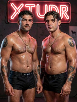 Two muscular men pose standing in front of a dark background with reddish lighting, both shirtless and wearing tight black boxers, showing shiny torsos, body hair, strong arms, and several visible tattoos on their shoulders, forearms, and torso. They wear gold chains around their necks and maintain a serious, intense expression, looking directly at the camera with a defiant attitude. Behind them stands out a red neon sign with the word “XTUDR,” creating a nocturnal, urban, and provocative atmosphere, reinforced by the shadows, the contrast of lights, and the studio photography aesthetic with a sensual and forceful tone.