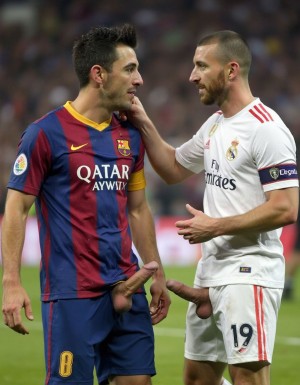 Dos futbolistas rivales conversan sobre el césped durante un partido, uno con la equipación azulgrana del FC Barcelona y patrocinio Qatar Airways, y el otro con la camiseta blanca del Real Madrid, brazalete de capitán y el número 19 en el pantalón. El jugador madridista, con barba y pelo rapado, apoya una mano en el hombro del barcelonista, que le mira de perfil con gesto serio. La escena tiene un tono llamativo y explícito porque ambos aparecen con el pene sobresaliendo por la parte delantera del pantalón corto, en una imagen que parece editada o manipulada, con el estadio desenfocado al fondo.