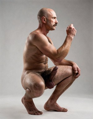 Un hombre desnudo, calvo y con bigote aparece en cuclillas de perfil sobre un fondo gris de estudio, con expresión seria y concentrada mientras mantiene una mano elevada con el puño suavemente cerrado. Su cuerpo velludo y musculoso muestra el torso, los brazos fuertes, el abdomen y la zona púbica, con el pene y el escroto visibles de forma frontal entre las piernas. La postura resalta una pierna apoyada en el suelo y una amputación en la otra extremidad, con el muñón flexionado cerca del cuerpo, transmitiendo una imagen corporal directa, íntima y escultórica, marcada por la desnudez, la fuerza física, la discapacidad y una composición sobria de retrato artístico.