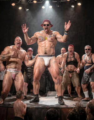 Un grupo de hombres musculosos y semidesnudos actúa sobre un escenario oscuro con focos teatrales, humo y público al frente, en una escena de espectáculo provocador tipo cabaret o performance fetichista. En el centro destaca un hombre alto con gorra roja, gafas de sol, botas negras y un suspensorio blanco ajustado que marca el pene, posando con los brazos levantados y la lengua fuera; lleva tatuajes en el pecho y una cruz dorada. A su alrededor aparecen otros hombres con arneses de cuero, slips, suspensorios, botas y accesorios rojos, algunos gritando o gesticulando con energía, creando una atmósfera intensa, sudorosa y exhibicionista de fiesta gay, burlesque y cultura leather.
