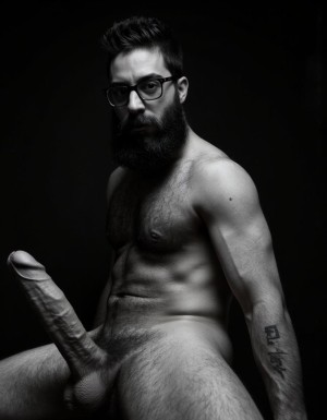 Fotografía hiperrealista en blanco y negro de un hombre adulto musculoso a la vista desde las rodillas para arriba hasta la cabeza, cuerpo muy delgado con musculación marcada y definida, rostro masculino, totalmente desnudo con polla erecta de treinta centímetros de longitud y ocho centímetros de diámetro, tiene rasgos marcados duros expresivos, barba densa poblada tupida oscura estilo hipster cubriendo completamente mandíbula mentón mejillas con grosor uniforme textura abundante muy definida, bigote denso tupido oscuro conectado perfectamente con la barba completa cubriendo completamente el labio superior, pelo oscuro medio peinado completamente recto hacia arriba con volumen considerable estilo hipster moderno quiff puntas verticales texturizadas hacia el cielo con altura prominente, cejas gruesas oscuras pobladas muy marcadas definidas, gafas de montura completa negra cuadradas grandes estilo hipster moderno sobre el puente de la nariz enmarcando unos ojos oscuros intensos que miran directamente a cámara con expresión seria penetrante confiada, expresión facial seria masculina sin sonrisa con mirada directa desafiante, pómulos marcados definidos angulares, nariz recta proporcionada, labios medios apenas visibles completamente cubiertos por bigote denso que conecta con la barba hipster completa, torso desnudo muy musculoso con pectorales enormes desarrollados y definidos, hombros anchos musculosos deltoide prominentes, brazos musculosos gruesos con bíceps y tríceps marcados visibles a ambos lados del cuerpo, abdomen tonificado marcado visible desde la cintura, piel con textura realista detallada poros imperfecciones sutiles, vello corporal denso oscuro abundante cubriendo pectorales abdomen brazos hombros con textura natural masculina muy visible, tatuaje en tinta negra en el bíceps del brazo derecho con la palabra exacta 