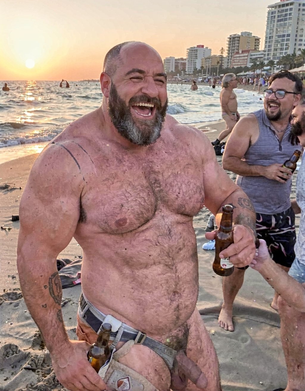 A naked, stocky, very muscular man laughs out loud on a beach at sunset while holding beer bottles in both hands; he wears only a kind of low belt or harness on his hips from which objects hang, leaving his chest hair, abdomen, arm tattoos, and penis visible. The scene has a festive and carefree atmosphere, with wet sand, waves, bathers in the sea, and several people around talking and drinking. In the background, tall buildings can be seen along the coast, palm trees, and a warm sunset illuminating the water and reinforcing the feeling of a beach party.